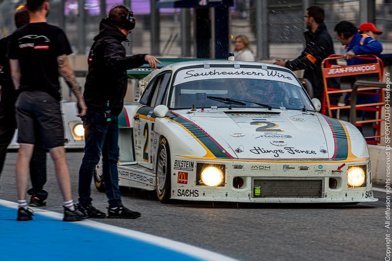 Voiture de course blanche avec des détails colorés sur la piste, entourée de mécaniciens et de spectateurs.